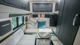 Cozy RV rental interior: cream diamond-stitched seating, dark overhead storage, wood floors, and a bright window view.