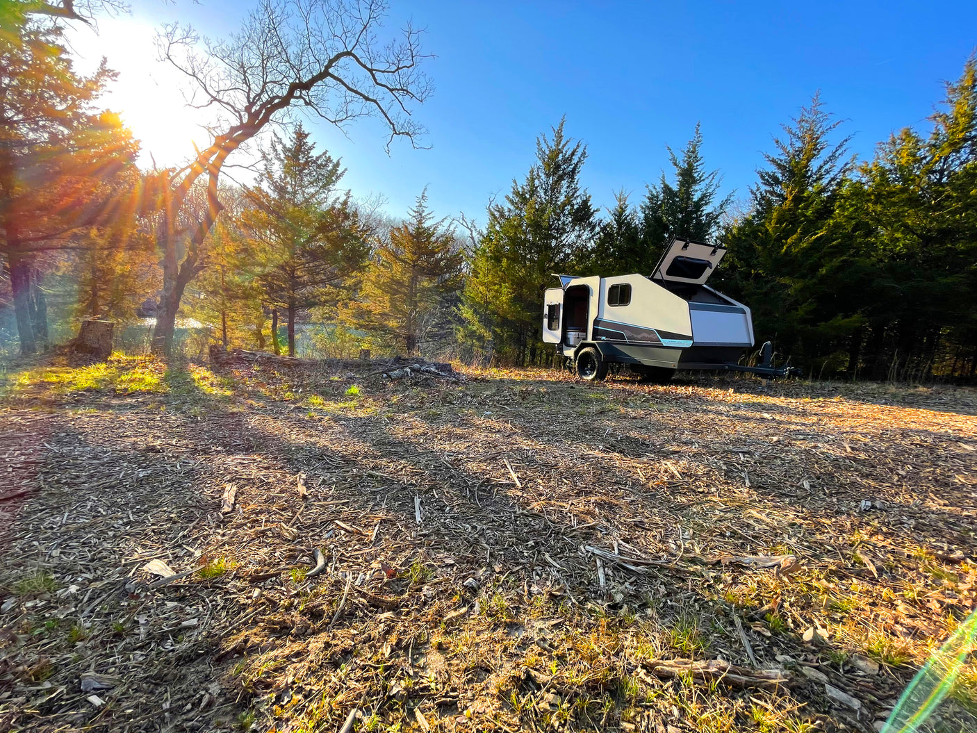 Sunny outdoor scene: A sleek camper trailer (RV rental) with open hatches, parked amidst trees and mulched ground.