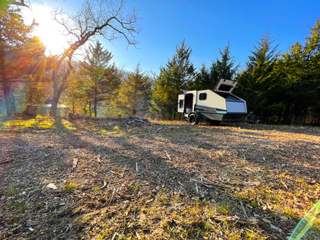 Sunny outdoor scene: A sleek camper trailer (RV rental) with open hatches, parked amidst trees and mulched ground.