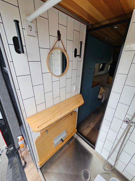 View of an RV rental's compact, modern bathroom area with white tiled walls, a fold-down counter, and a steel shower pan.