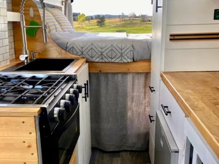 Inside an RV rental: a compact kitchen with stove/oven, sink, and a cozy elevated bed with a view. — photo 7