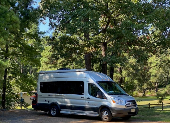 Experience nature with this silver camper van RV rental, parked at a sunny wooded campsite.