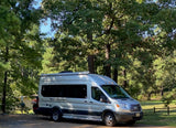 Experience nature with this silver camper van RV rental, parked at a sunny wooded campsite.