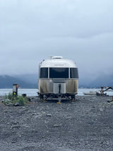 A silver Airstream rv rental rests on a rocky shore by the water, surrounded by misty mountains on an overcast day.