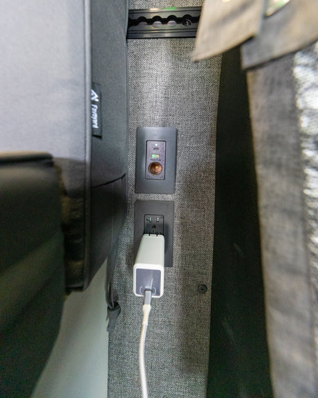 Modern RV rental charging hub: USB-C, USB-A, 12V, and AC outlet with a charger plugged into a gray textured wall.