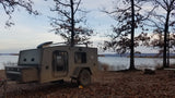 An Escapade Camper Co. teardrop trailer (RV rental) is set up for lakeside camping, surrounded by fall trees and leaves.