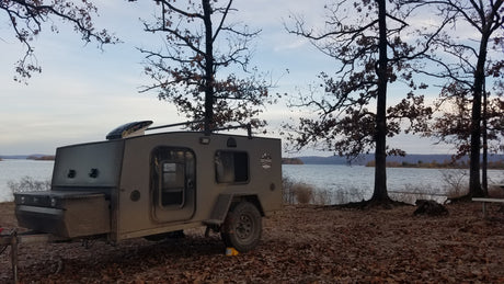 An Escapade Camper Co. teardrop trailer (RV rental) is set up for lakeside camping, surrounded by fall trees and leaves.
