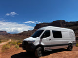 White rv rental Sprinter van parked on a dirt road in a vast desert with towering red cliffs under a blue sky.