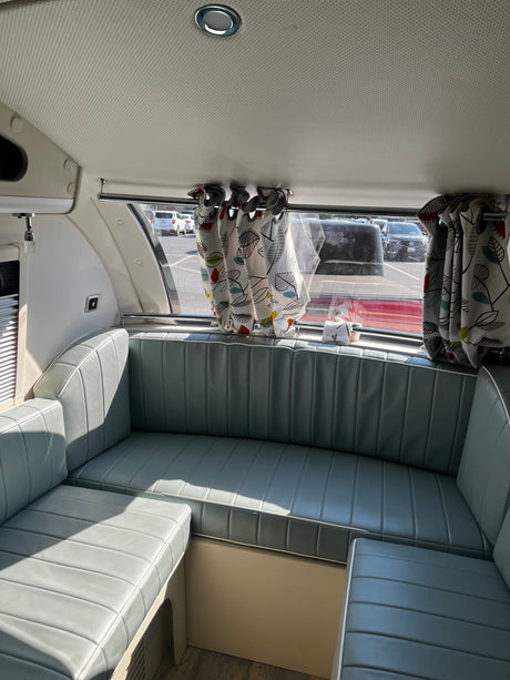 Relaxing RV rental interior with light blue cushioned seating, artistic curtains, and sunny window view.