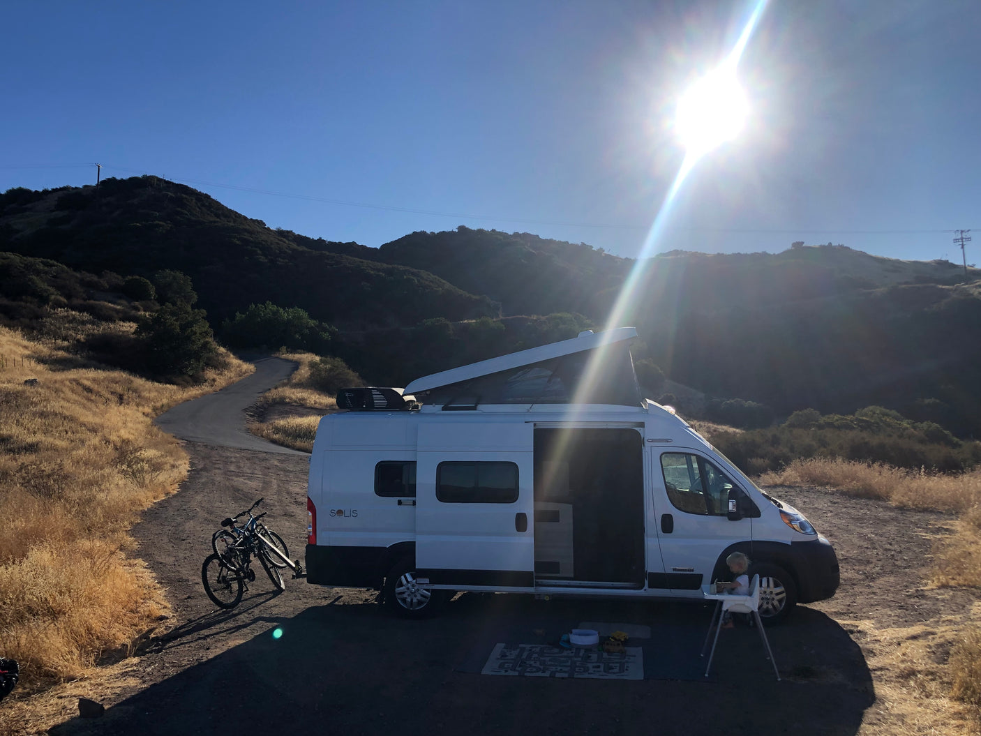An rv rental, a Winnebago Solis, parked in a sunny mountain landscape. Bikes attached, child plays by the open door.