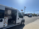 A white camper van, likely an RV rental, with its side door open, parked by a motel and distant mountains on a sunny day.