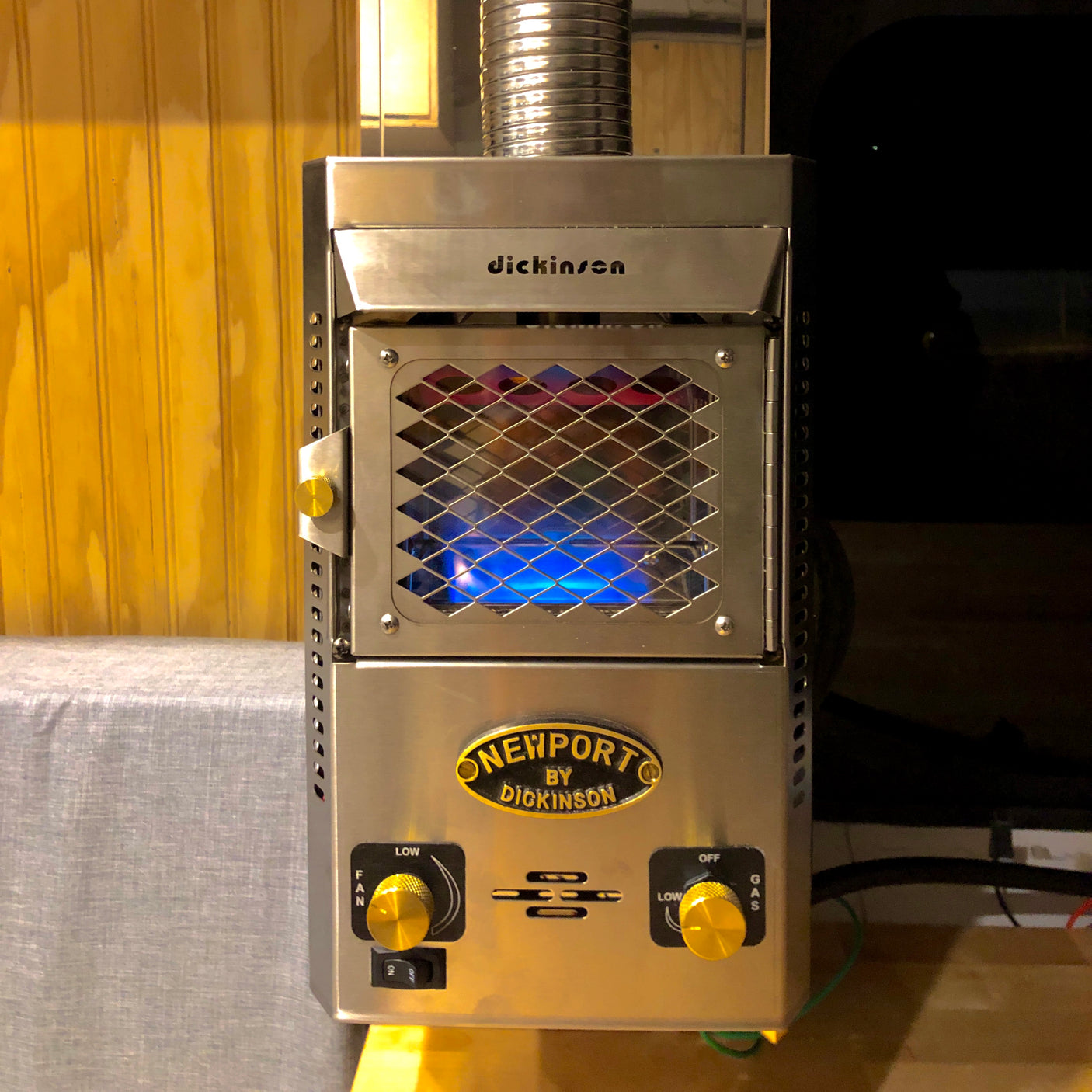 A stainless steel Dickinson Newport propane heater with active blue flames, ideal for an RV rental. Features control knobs and a vent pipe.