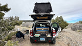 White SUV RV rental setup for car camping, featuring a rooftop tent & rear camp kitchen. Desert scene with person relaxing under awning.
