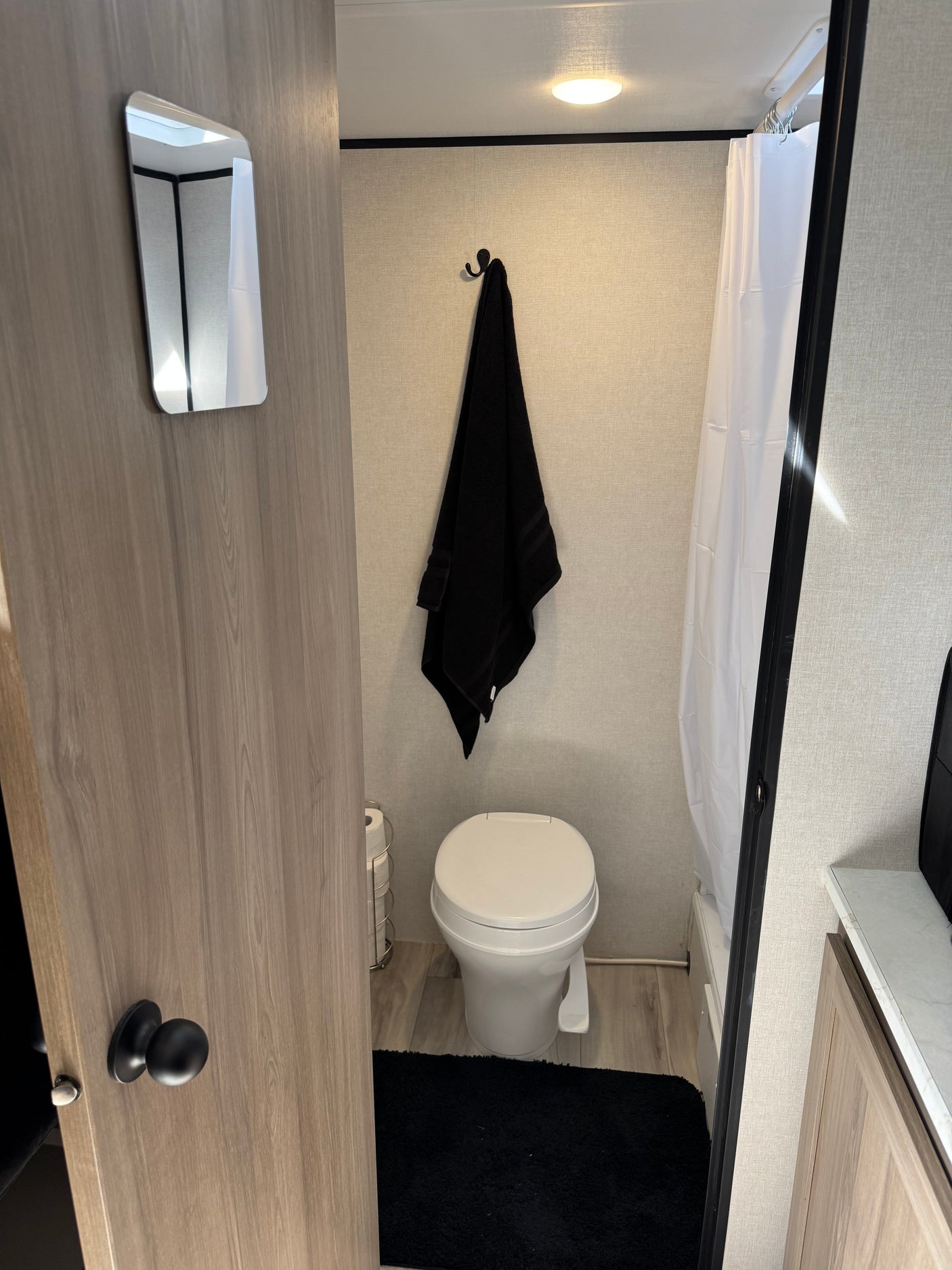 Inside an RV rental bathroom: a white toilet, black towel, shower curtain, and a mirror on the door. Compact and functional.