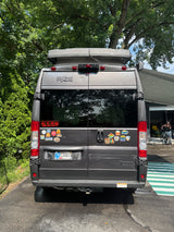 Rear view of a gray **RV rental**, a camper van with a pop-top roof and adventure stickers, parked outdoors.
