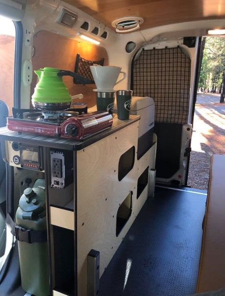 RV rental interior: a compact kitchen setup with a stove, kettle, coffee gear, fridge, and storage. Ideal for van life.