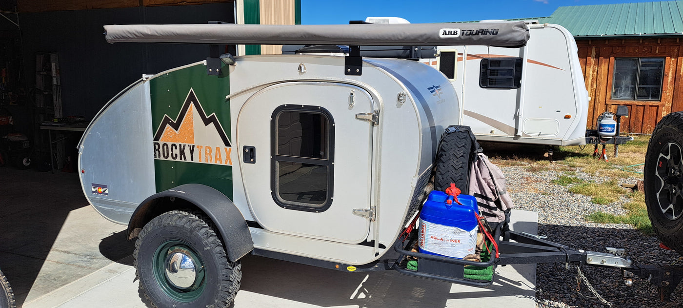 This teardrop RV rental features an ARB awning and adventure gear. A larger travel trailer and building are in the background. — photo 9