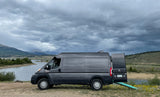 A gray RV rental camper van parked by a serene lake under stormy clouds, with mountains in the background. Ideal for adventure.