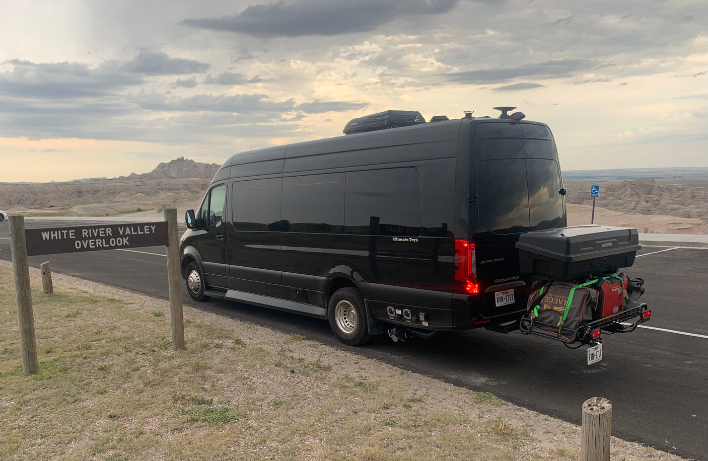 A black luxury rv rental van at White River Valley Overlook, with a rugged landscape under a cloudy sky.