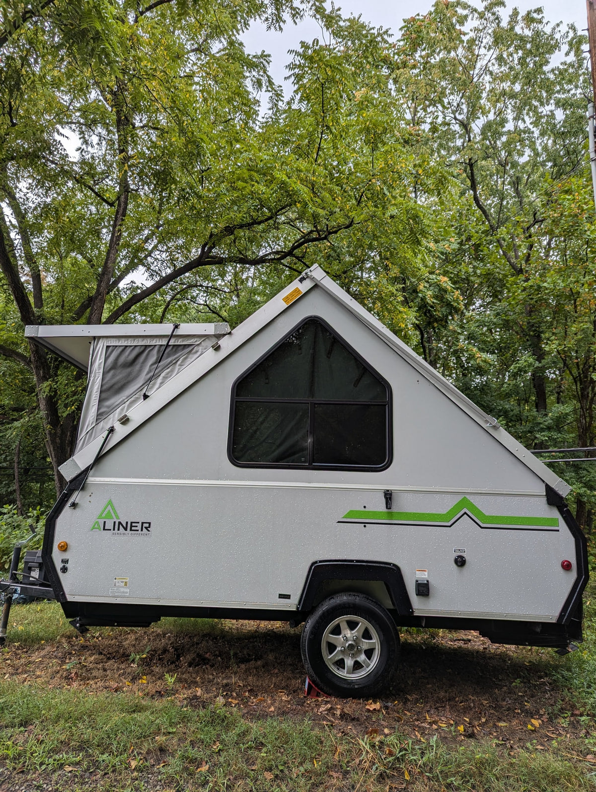 A gray A-liner A-frame pop-up camper, suitable for rv rental, parked in a lush, tree-filled setting with rain droplets visible. — photo 2