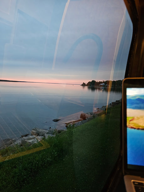 Serene lake sunset viewed from an RV rental window, showing interior reflections and a laptop. Ideal for scenic travel.