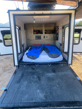 Inside a compact rv rental: a bed with two sleeping bags, overhead storage, and a lowered rear ramp for easy access.