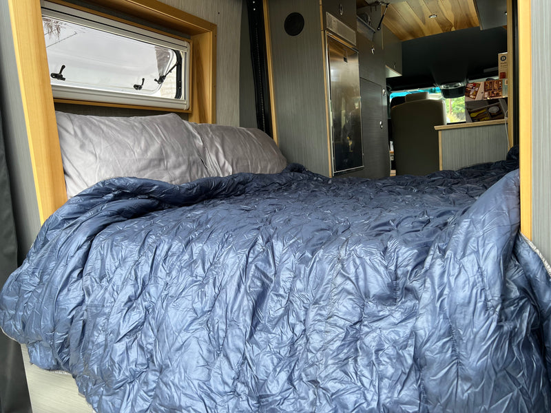 Inside a modern RV rental: a comfortable bed with a blue comforter and gray pillow, with a window and kitchen visible. — photo 13
