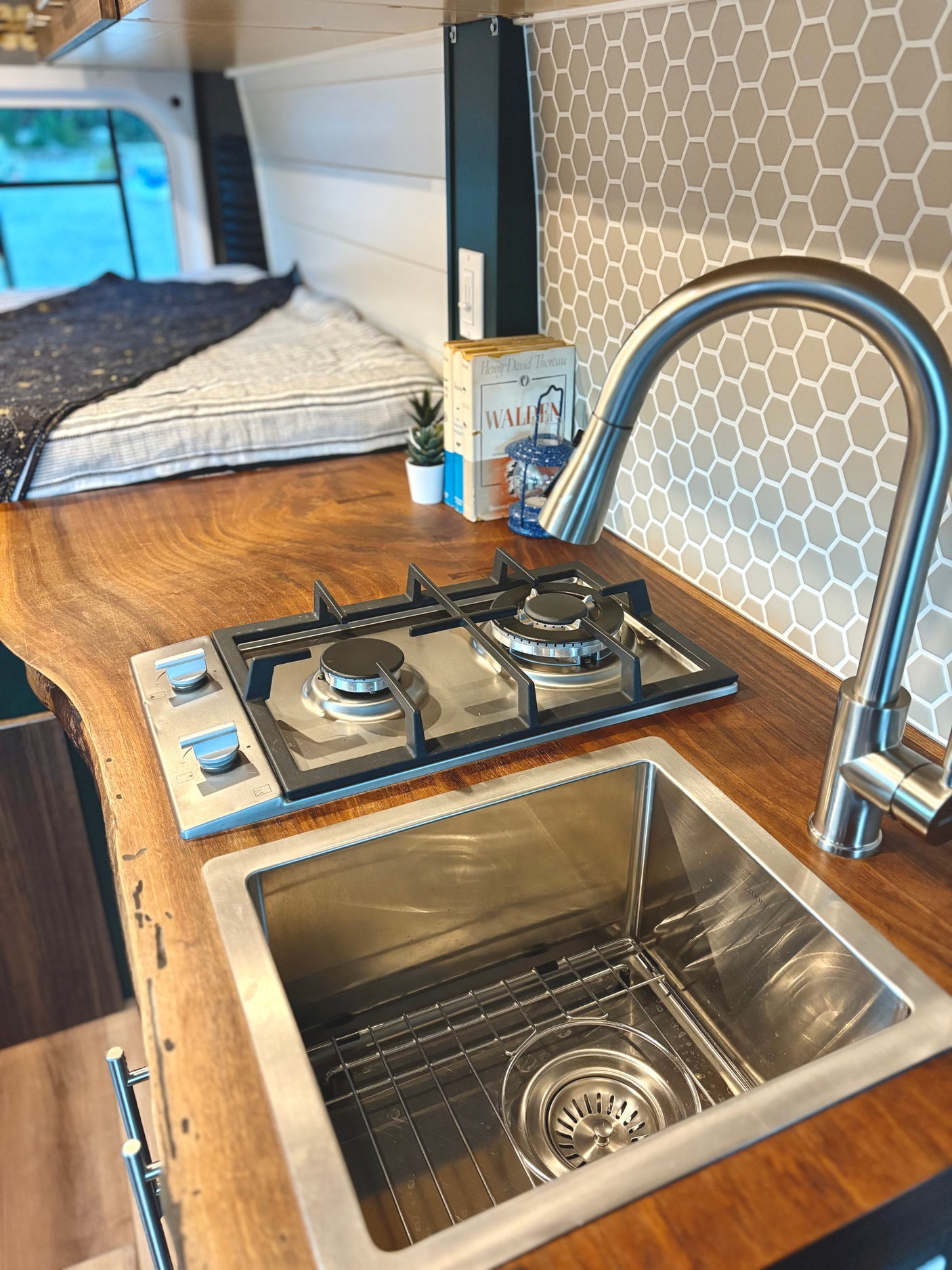 RV rental kitchen: live edge wood counter with sink, stove, and hex tile backsplash in a cozy camper van.