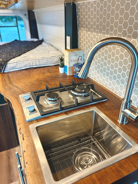 RV rental kitchen: live edge wood counter with sink, stove, and hex tile backsplash in a cozy camper van.