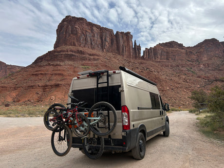 An adventure-ready RV rental with bikes attached, set against majestic red rock formations under a cloudy sky.