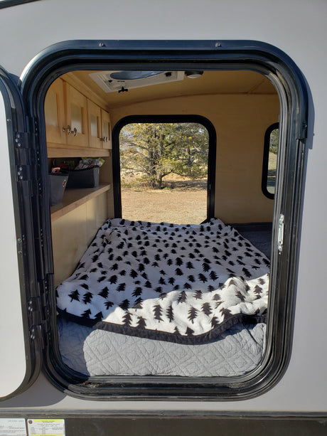 Cozy rv rental interior: A compact camper with a bed, tree-patterned blanket, cabinets, and a window view of nature.