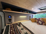 Inside a comfortable RV rental: bed, controls, storage, and a scenic forest view through the window.