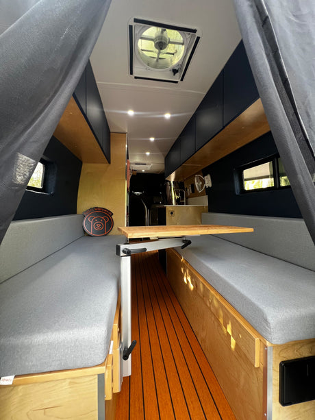 Modern RV rental interior: convertible dinette with gray cushions, wooden floor, overhead storage, and ceiling fan.