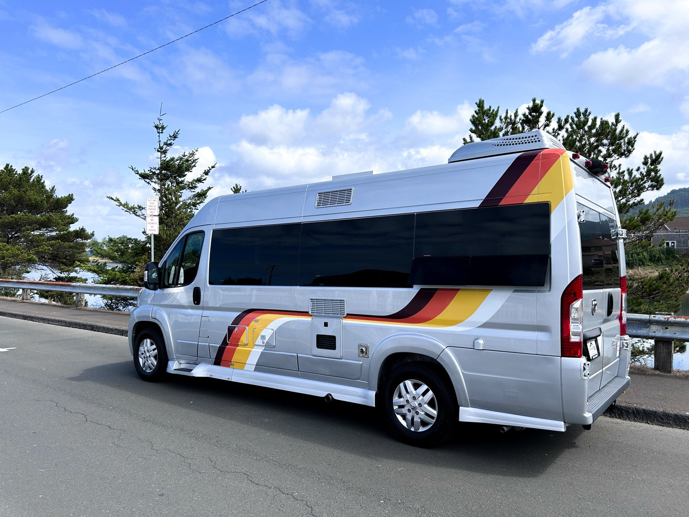 parked by a scenic bay. Note the 'no overnight parking' sign for RVs.|Silver RV rental with colorful stripes parked on a gravel lot amidst green trees. This camper van is ready for your adventure.|Mobile office setup in an RV rental: laptop on a fold-down desk — photo 3