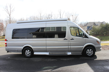 A silver Mercedes-Benz Sprinter camper van, ideal for RV rental. Seen from the side, parked on asphalt with houses and trees.