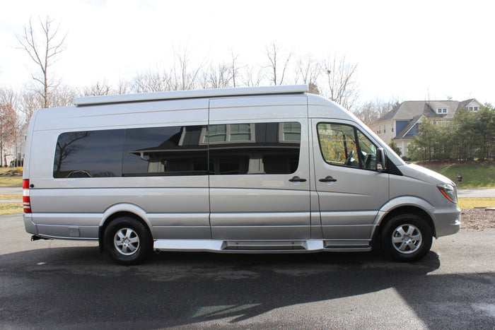 A silver Mercedes-Benz Sprinter camper van, ideal for RV rental. Seen from the side, parked on asphalt with houses and trees.