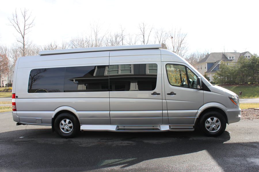 A silver Mercedes-Benz Sprinter camper van, ideal for RV rental. Seen from the side, parked on asphalt with houses and trees.