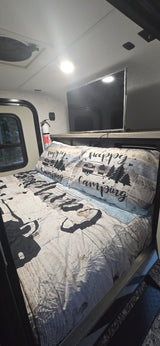 Cozy RV rental bedroom featuring 'Happy Camping' themed bedding, a TV, and a window offering a view of outdoor greenery.