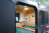 Cozy RV rental interior shows a colorful bed, wooden walls, and string lights, viewed from the open doorway.