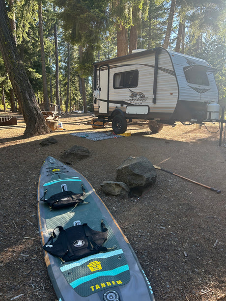 An RV rental is parked among towering trees at a campsite, with a paddleboard and life vest ready for water fun.