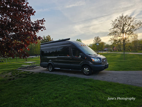 RV rental: Dark grey campervan parked by a picnic table in a lush park, sun setting through trees.