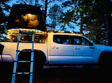 White pickup truck with illuminated rooftop tent, open, ladder extended. Great for overlanding or an RV rental adventure.