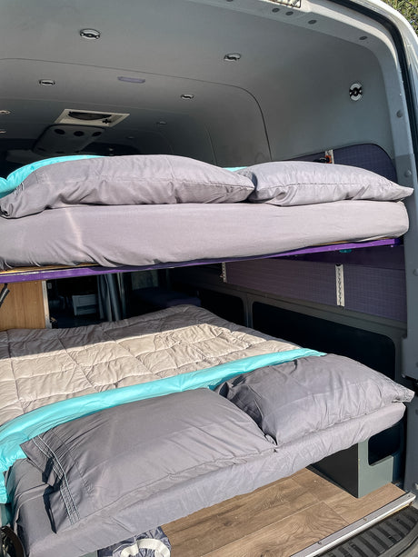 Inside an RV rental, two comfortable bunk beds are made with grey and turquoise bedding, ready for travelers.