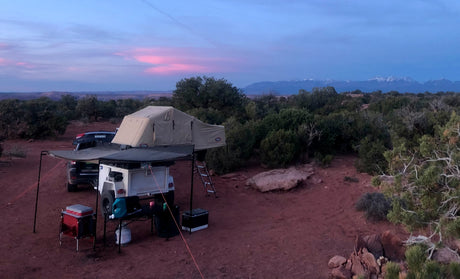 An overland rv rental setup: rooftop tent on trailer, SUV, & gear in a desert landscape at dusk.