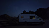White camper van RV rental parked at night with illuminated interior, under a clear, starry sky & dark rocky terrain.
