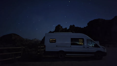 White camper van RV rental parked at night with illuminated interior, under a clear, starry sky & dark rocky terrain.