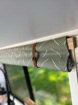 Stylish RV rental window blind: green fabric with white pattern, rolled up and secured by a brown leather strap.