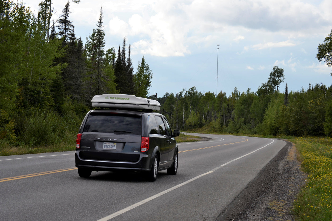A minivan RV rental with 'The Penthouse' rooftop tent drives on a scenic road through a forest, with a cell tower in the distance.