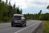 A minivan RV rental with 'The Penthouse' rooftop tent drives on a scenic road through a forest, with a cell tower in the distance.