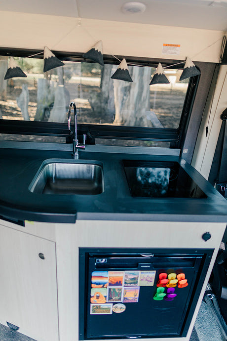 Compact kitchenette in an RV rental, featuring a sink, stovetop, and a fridge adorned with travel magnets. Ideal for van life.